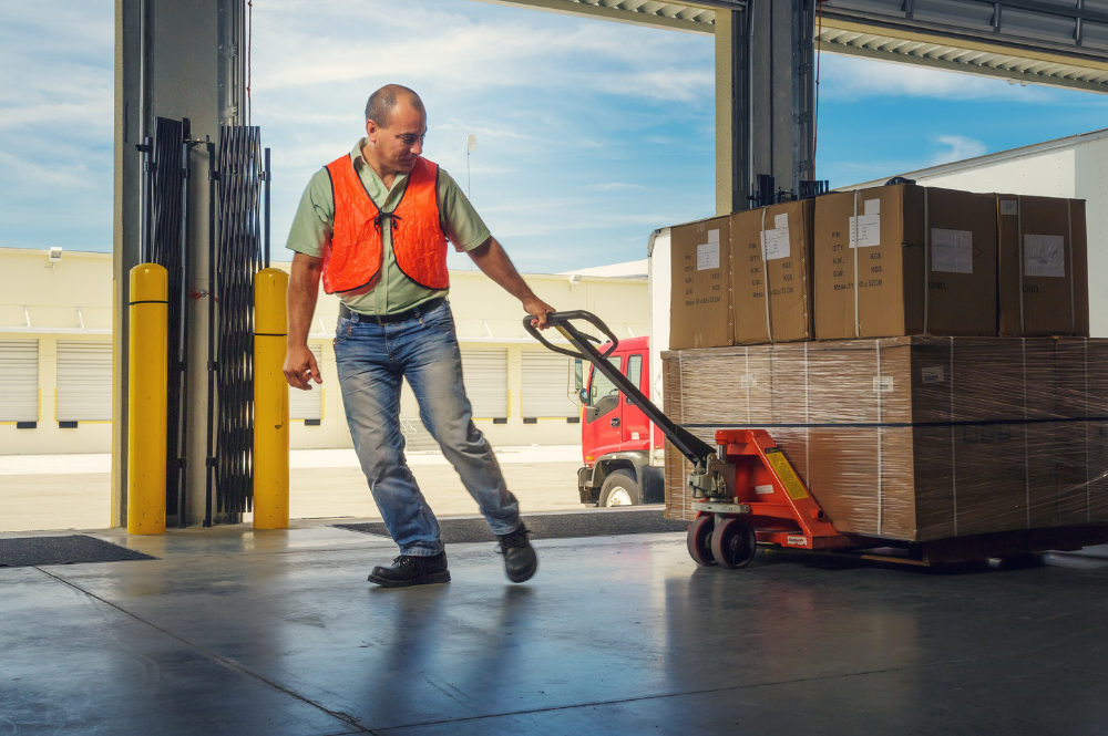 A man in an orange vest pulls pallets loaded with boxes intended for manufacturing.